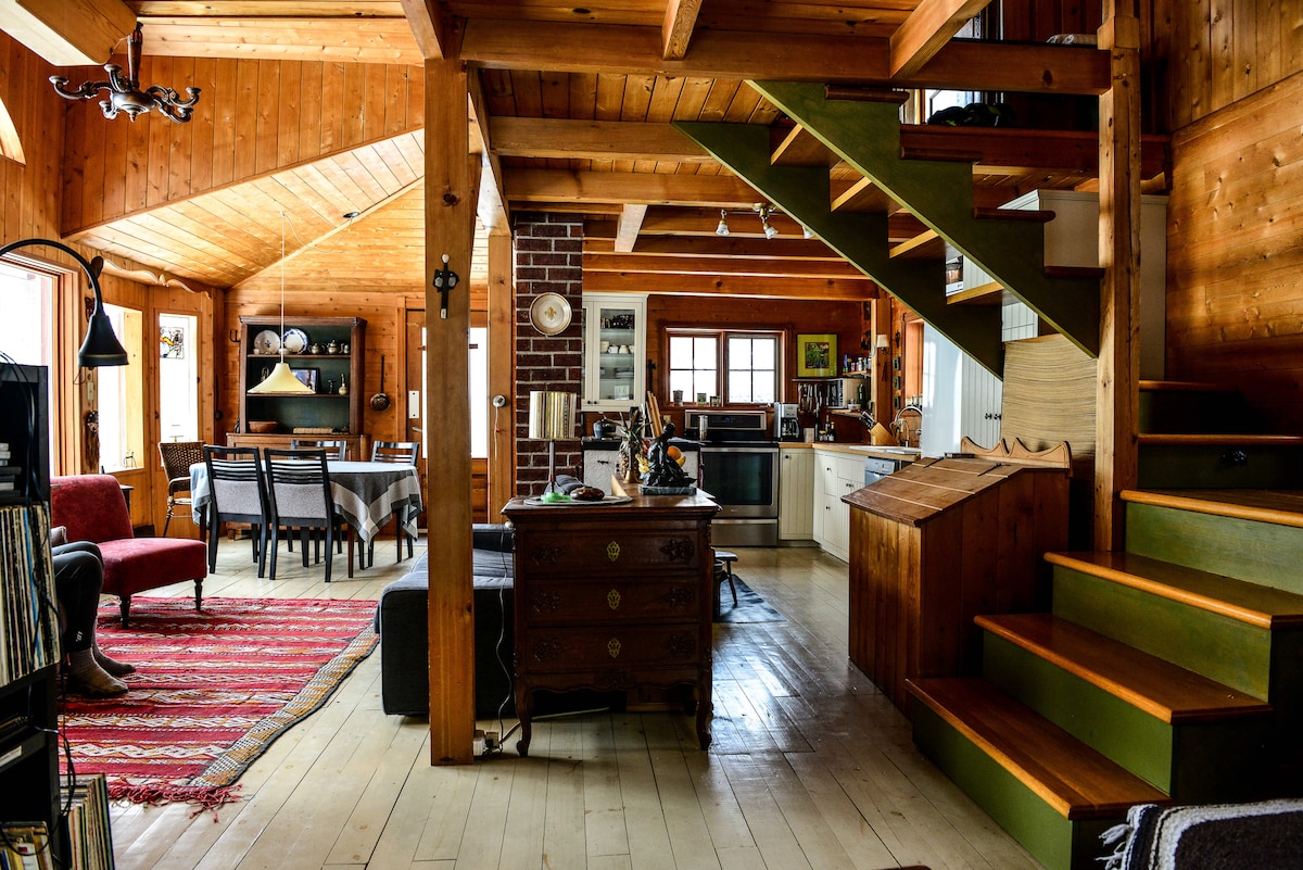 A warm interior showcases natural wood finishes and a staircase leading to the upper level. A dining table is set in the center, surrounded by cozy seating options. A kitchen area is visible at the back, complemented by decorative elements that enhance the space.