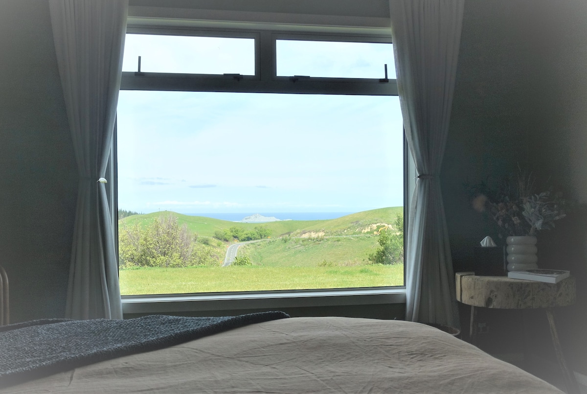 A spacious bedroom is set with a view through large windows overlooking lush green hills leading to the coastline. Soft curtains frame the scene, while a comfortable bed is positioned prominently in the foreground.