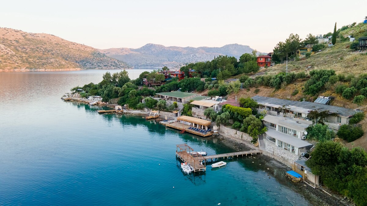 The shoreline is lined with elegant accommodations, nestled among lush greenery. Private docks extend into the calm blue waters, offering direct access to the sea. Rolling hills create a serene backdrop, enhancing the peaceful setting of this beachfront location.