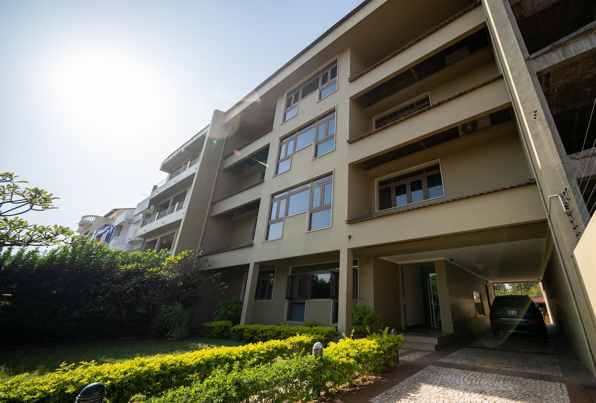 The modern apartment building features multiple floors with large windows that allow natural light. A green landscaped area with shrubs lines the front, and a covered parking space is visible on the right side, providing convenient access for residents.