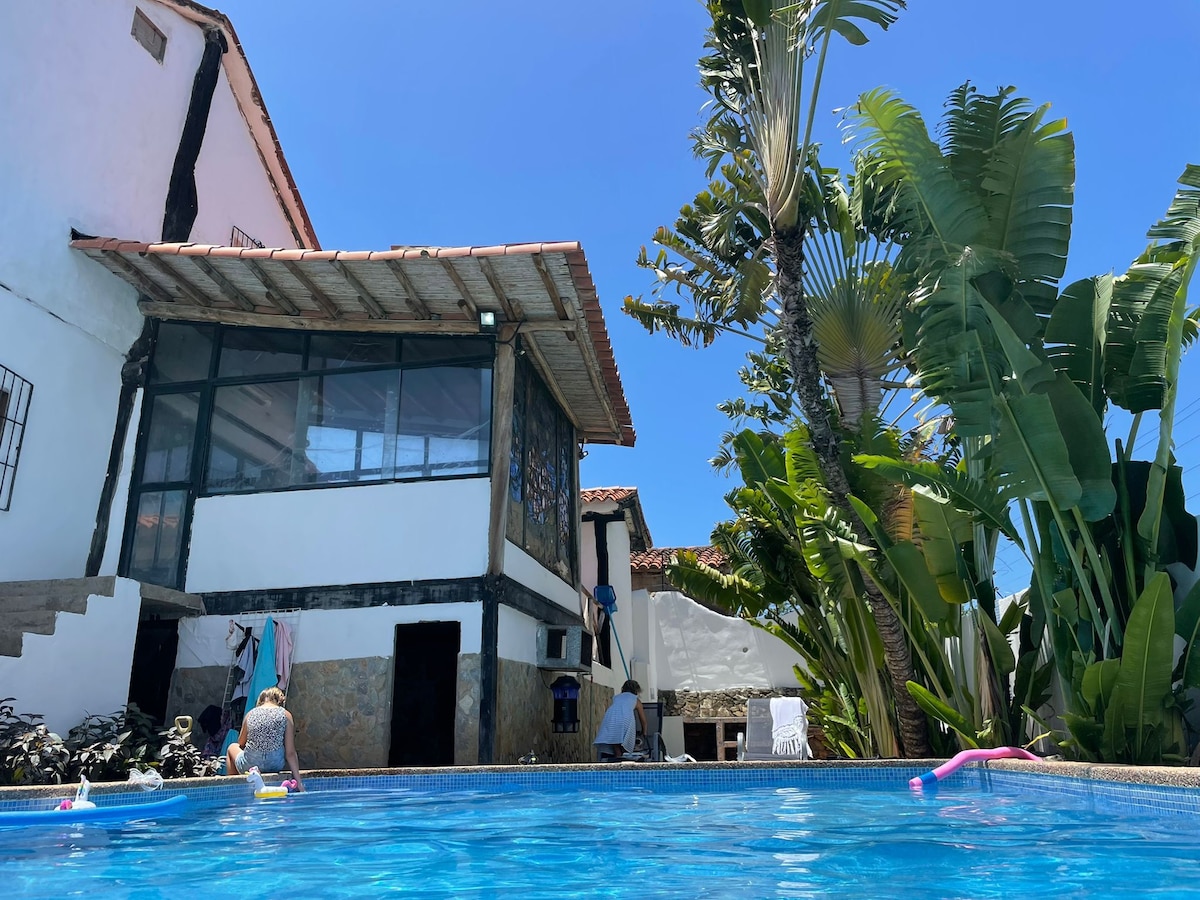 A private villa is set against a clear blue sky, featuring a swimming pool surrounded by lush greenery and palm trees. The house is partially obscured by foliage, displaying a mix of traditional and modern architecture with large windows. Sun loungers are visible near the pool.