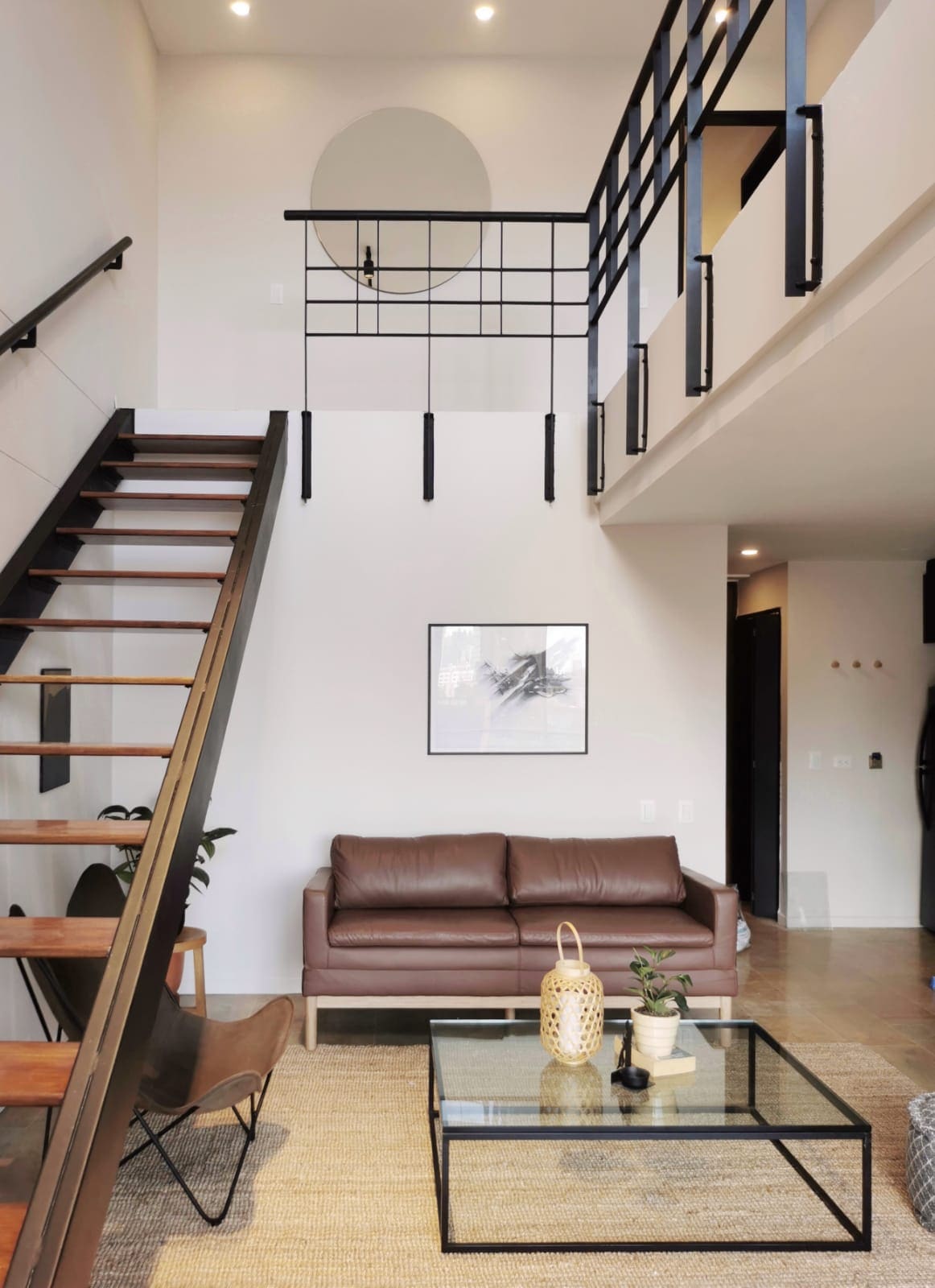 The open living area features a brown leather sofa positioned next to a coffee table with a decorative plant and a stylish lantern. A staircase leads to the upper level, where a minimalist railing separates the space, while artwork adorns the wall.