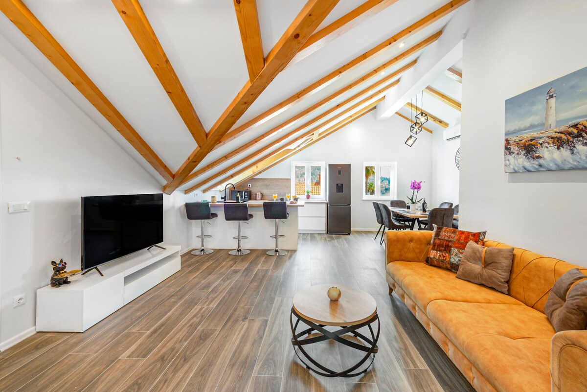 An open-plan living area features a spacious orange sofa and a round coffee table at the center. A modern kitchen with bar seating and stainless steel appliances is visible. Exposed wooden beams add character to the high ceiling, while natural light fills the room.