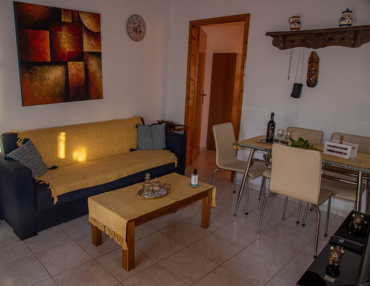 A living area is depicted, featuring a sofa with a yellow throw, a wooden coffee table, and a dining table with two chairs. Wall decor includes an abstract painting and a carved wooden mask. Natural light brightens the tiled floor.