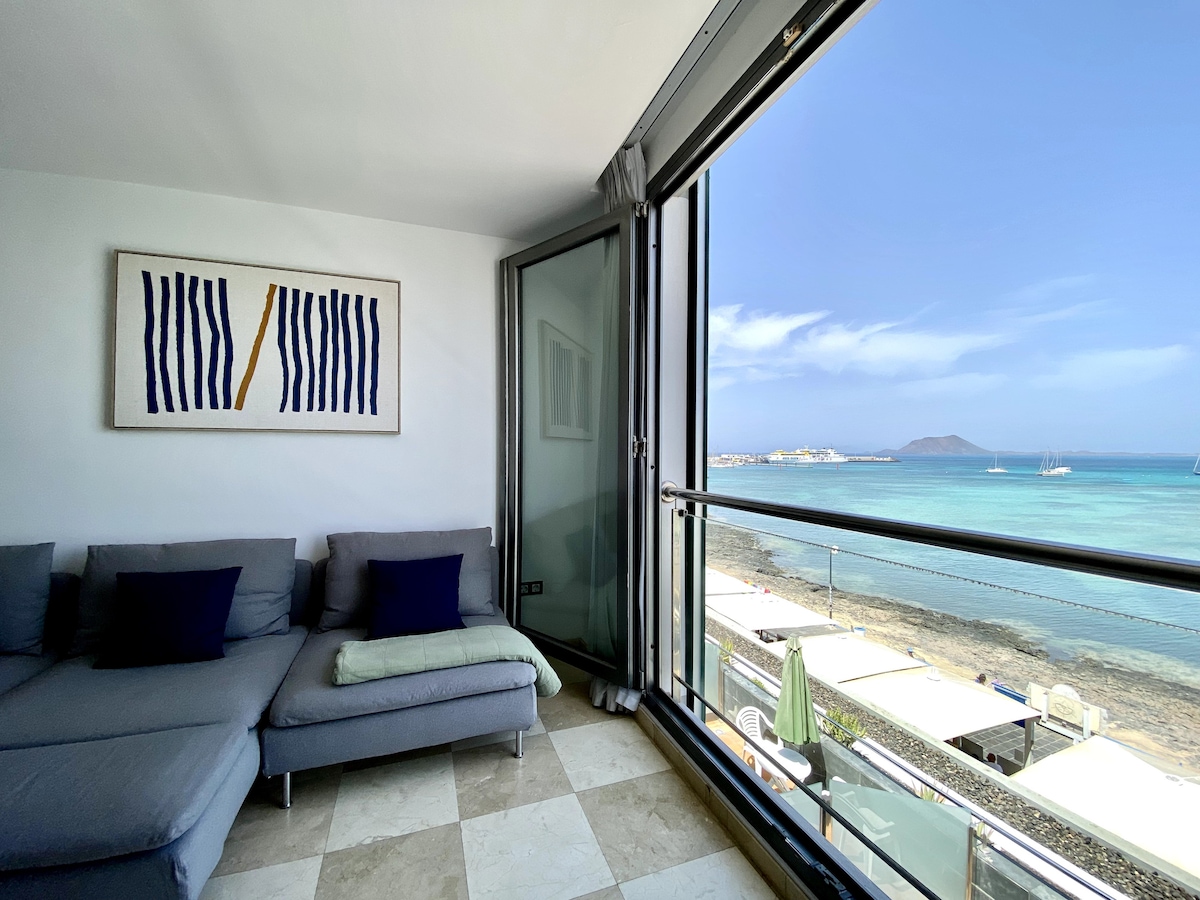 A cozy seating area is featured, with a gray sectional sofa adorned with blue cushions. Floor-to-ceiling glass doors open to reveal scenic views of the bay and the island of Lobos, allowing abundant natural light into the space.