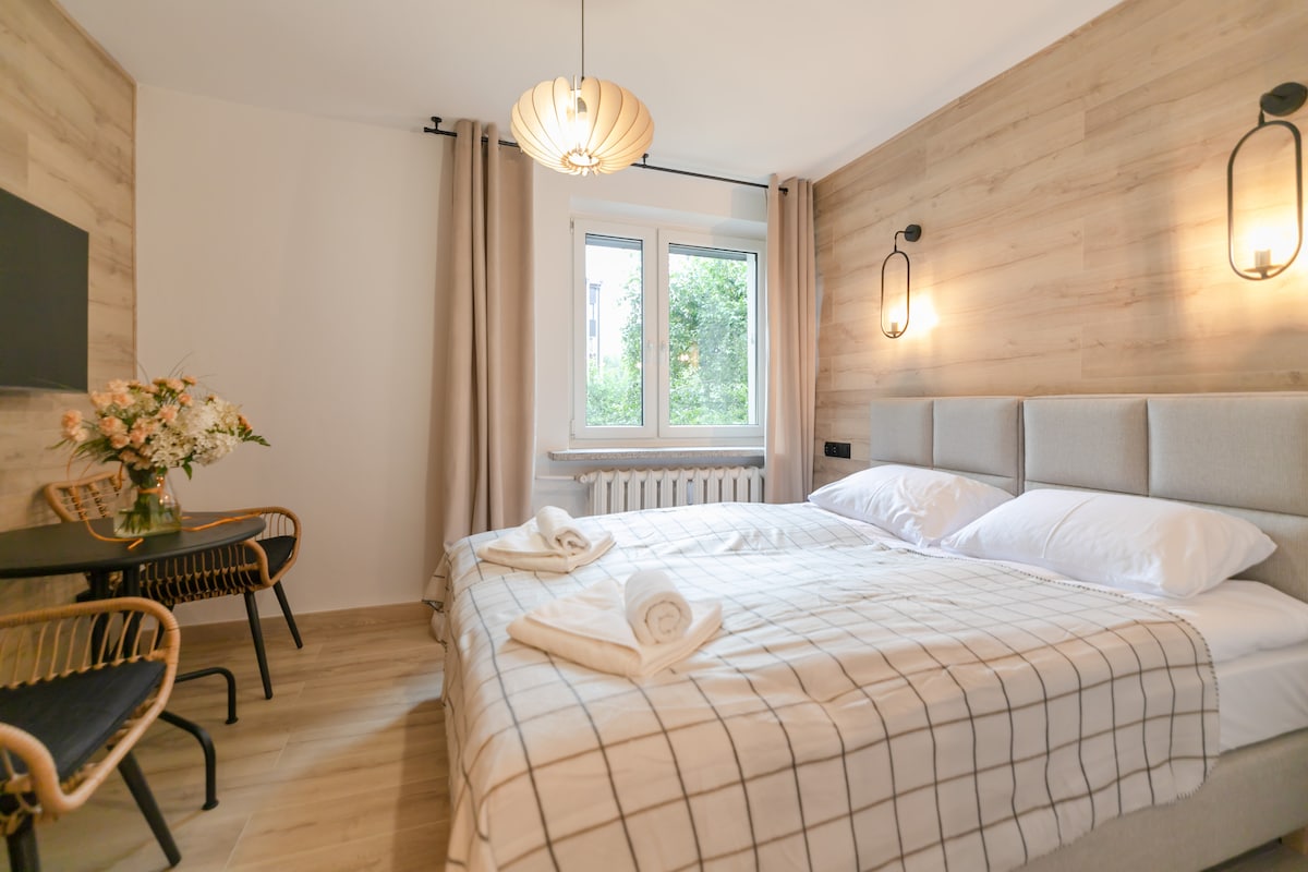 A well-appointed bedroom featuring a spacious bed with neatly arranged linens. Soft, neutral tones are accented by warm lighting fixtures. A small dining area is present, complete with a wicker chair and a simple table. Natural light filters in through the window, framed by light curtains.