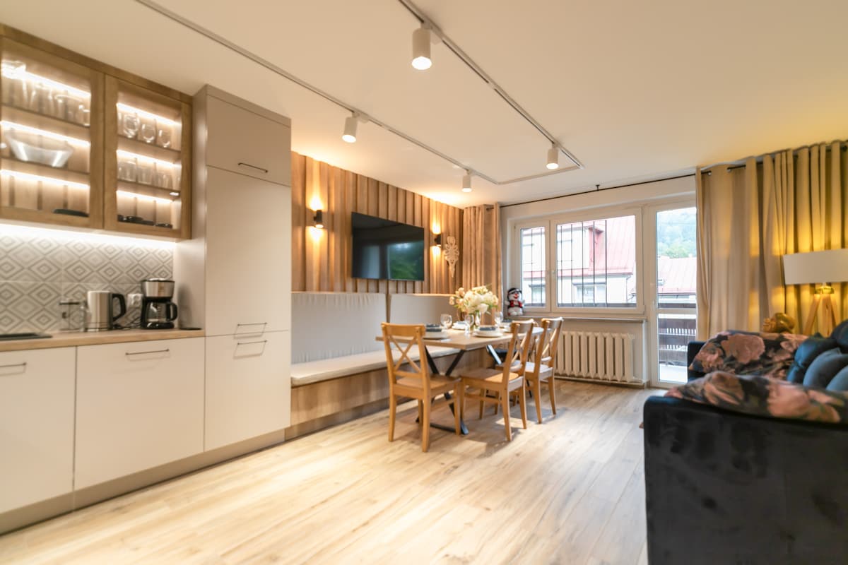 A modern kitchen area is equipped with a coffee maker and glass cabinetry, complemented by a dining table surrounded by four wooden chairs. Soft lighting highlights the elegant wooden paneling, while a cozy seating area with floral accents offers comfort. A window provides natural light and outdoor views.