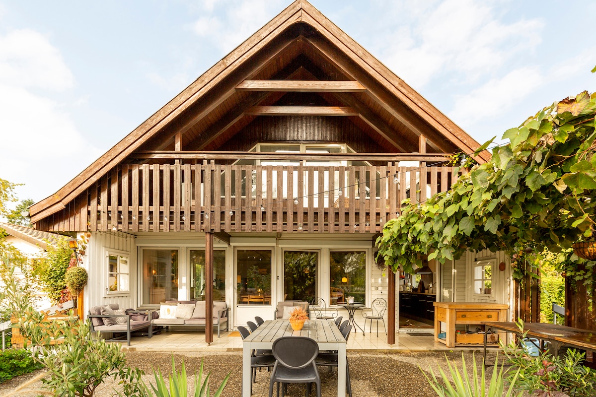 A welcoming exterior view of the apartment reveals a wooden structure with a high, sloped roof. A terrace with outdoor seating and a dining table is visible below, surrounded by greenery. Large windows allow natural light into the living space.