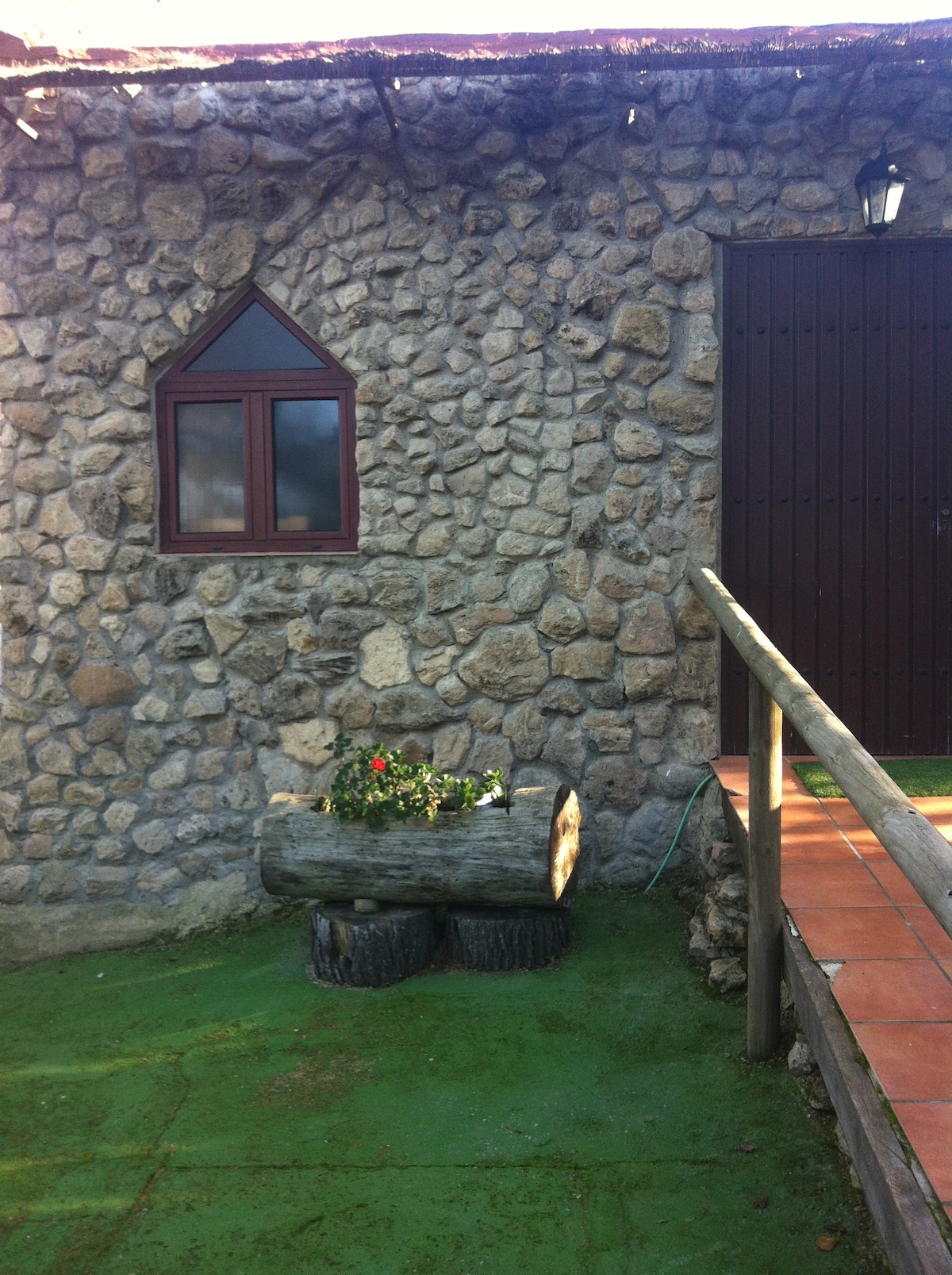 A stone exterior of a rustic building features a distinct arched window with a wooden frame. Below, a decorative planter made from logs holds vibrant flowers. A wooden railing leads to the entrance, adding to the inviting atmosphere of the outdoor space.