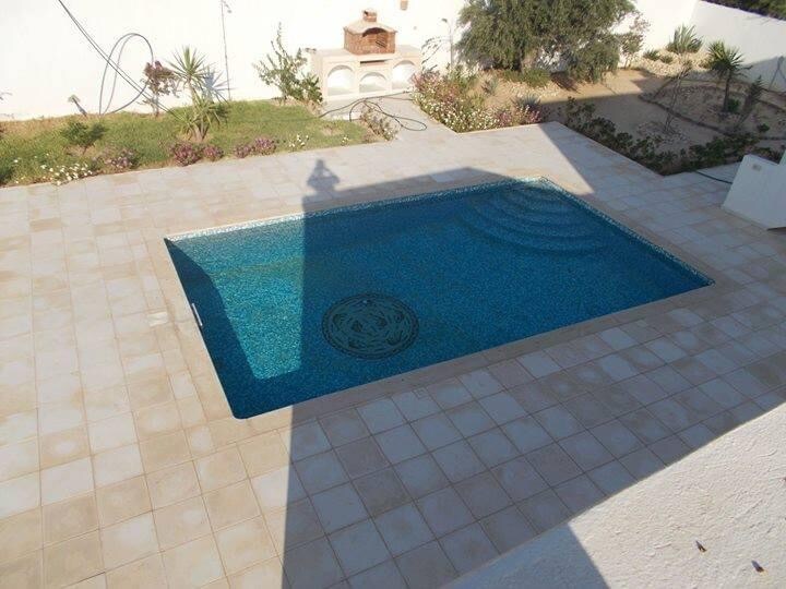 A rectangular swimming pool is surrounded by light-colored tiled flooring. The water appears clear, reflecting the sunlight, with gentle ripples visible on the surface. Lush greenery lines the pool area, contributing to a serene outdoor setting.