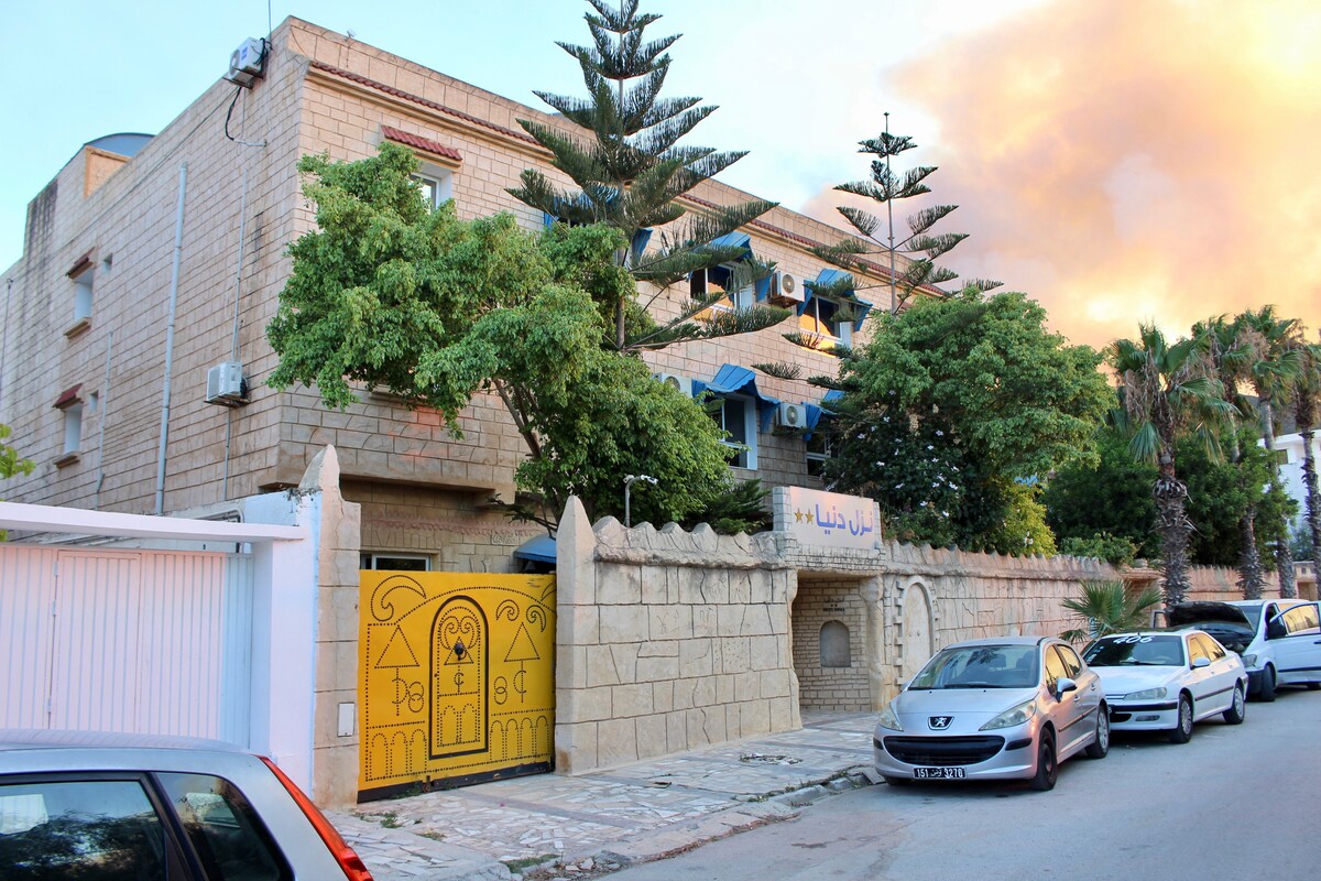 The exterior of Hotel Donia is shown, featuring a stone façade with large windows. Lush trees and palms are seen surrounding the entrance, which is marked by a vibrant yellow gate. Vehicles are parked along the street, adding to the lively setting.