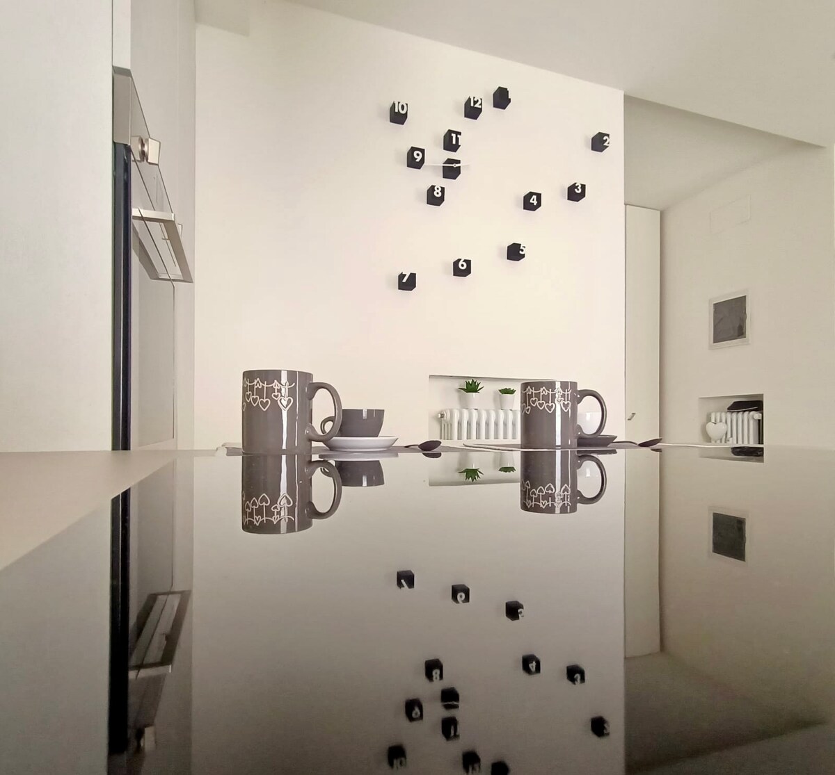 A modern kitchen space is featured, showcasing sleek cabinetry and a glossy countertop. Two coffee mugs and matching plates are arranged on the counter. A unique wall display with small black shelves and a plant adds visual interest, reflecting softly on the smooth surface.