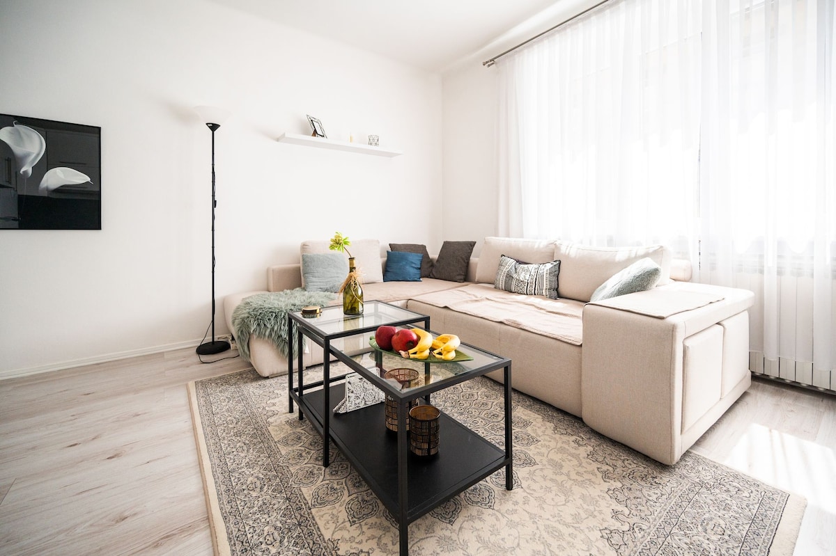 A spacious living area features a large light-colored sectional sofa with assorted decorative pillows. A glass coffee table sits in the center, adorned with fresh fruit and a small vase of flowers. Natural light filters through sheer curtains, adding warmth to the room.