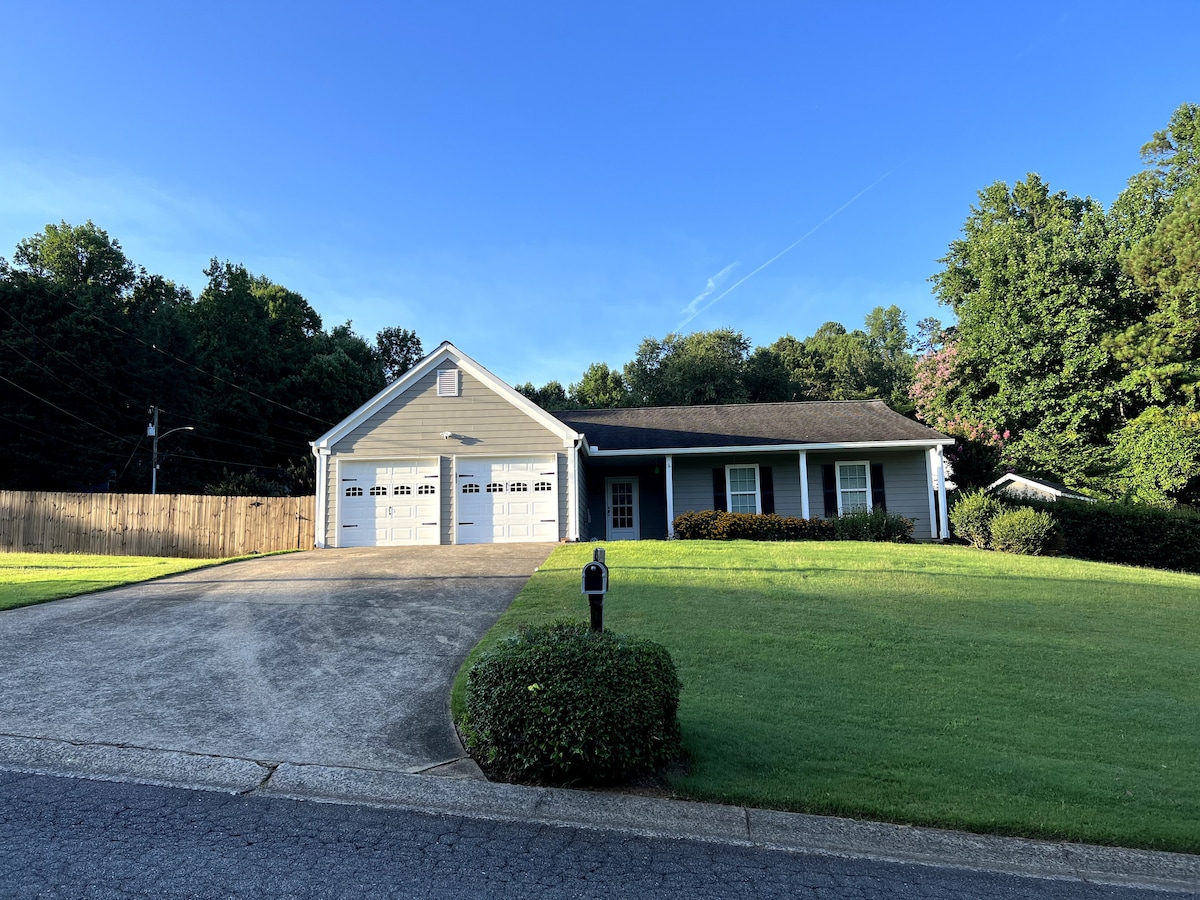 A single-story ranch-style home with a well-maintained exterior is depicted. The driveway leads to a garage with two doors, while a lawn surrounds the house. Greenery and trees are visible in the background, providing a serene setting.
