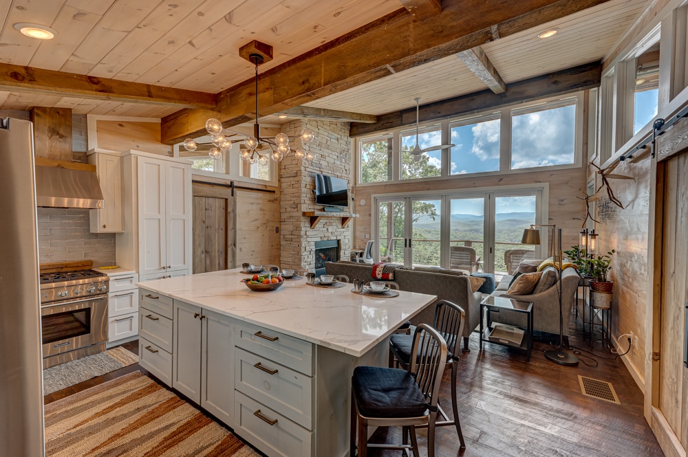 A modern kitchen is showcased, featuring a spacious island with a marble countertop. Large windows invite natural light, offering views of the surrounding landscape. Wooden beams run across the ceiling, complementing the stone accents and a cozy seating area nearby.