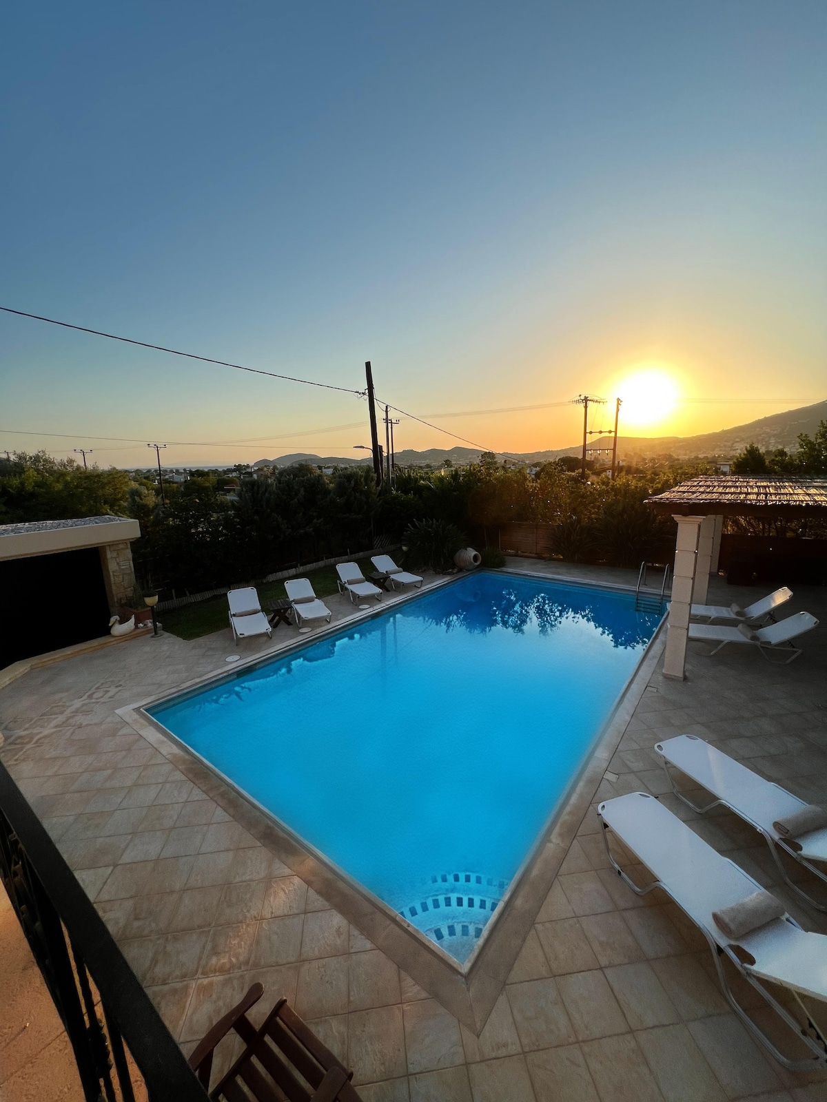 A large swimming pool is surrounded by sun loungers, reflecting the warm hues of the setting sun. The outdoor area features a covered patio and lush greenery, creating a serene environment. Mountain silhouettes frame the distant horizon, enhancing the tranquil setting.
