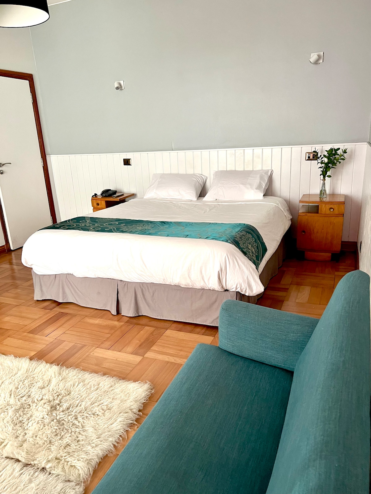 A cozy sleeping area is shown with a spacious bed dressed in white linens and a teal decorative throw. Natural wood flooring complements the room's neutral walls and a small green plant sits atop the bedside table. A soft area rug and a simple couch add to the inviting atmosphere.