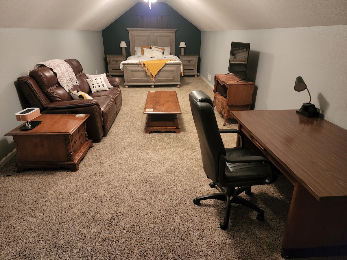 An open loft-style room features a spacious bed with a decorative yellow throw. A seating area consists of a leather loveseat and a wooden coffee table. A desk with a task lamp is situated next to a TV on a stand, providing a functional workspace.