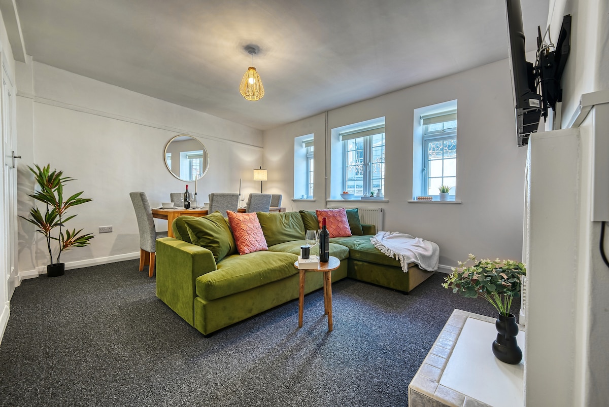 A cozy lounge area showcases a green sectional sofa adorned with decorative pillows. A round mirror and dining table with four chairs are visible, illuminated by natural light from windows. Decorative plants and a TV add to the welcoming ambiance of the space.