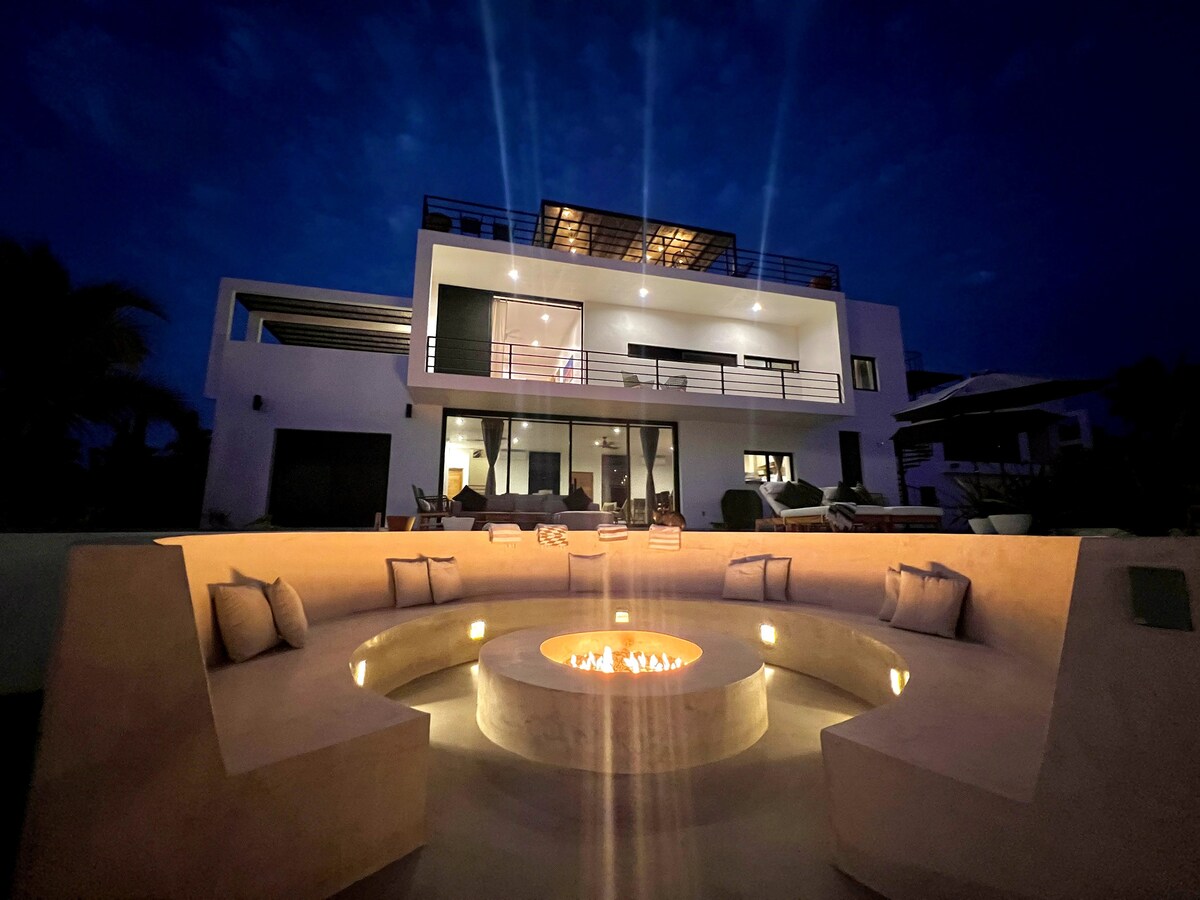 A modern exterior view of the house is illuminated at night, showcasing a spacious second-floor balcony and expansive glass doors. A circular seating area surrounds a glowing firepit, creating an inviting social space. Soft lighting highlights the contours of the architectural design.