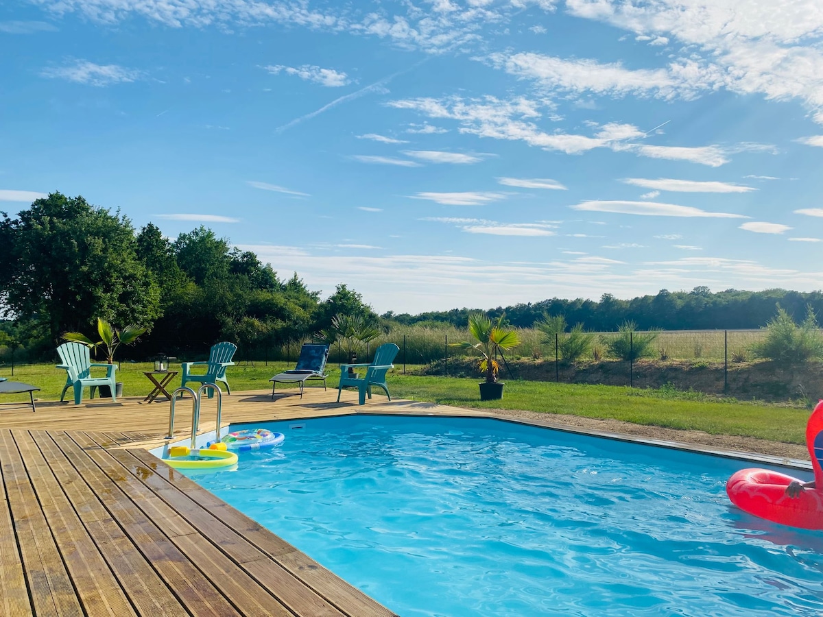 An outdoor swimming pool is surrounded by wooden decking, featuring bright lounge chairs in shades of green and blue. Lush greenery and open fields are visible in the background under a clear blue sky, enhancing the serene outdoor setting.
