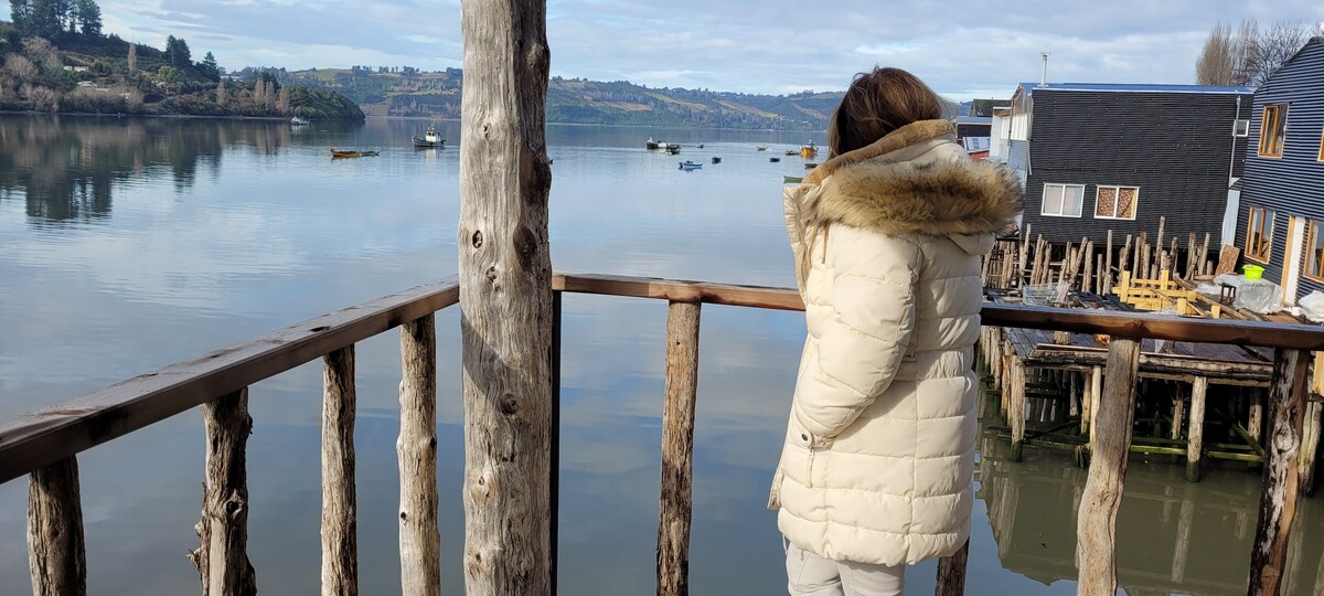A wooden balcony railing overlooks a calm body of water, reflecting the surrounding hills and boats. A person in a light-colored jacket stands near the edge, gazing at the view, which exhibits a tranquil scene of nature.