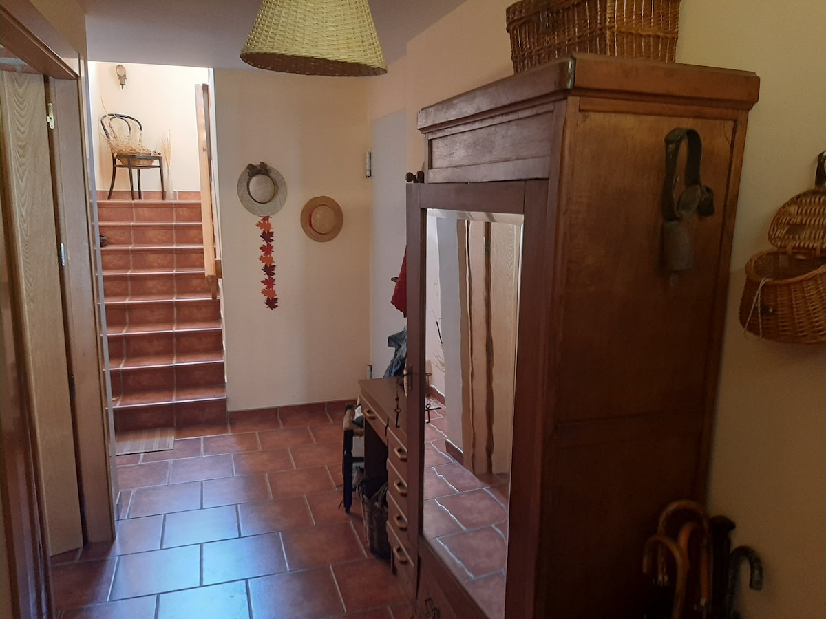 A welcoming entryway is featured, showcasing terracotta tiles and warm lighting. A wooden wardrobe with a mirror sits adjacent to a console table. Decorative baskets and hats hang on the wall, and a staircase with wooden steps leads to the upper level.