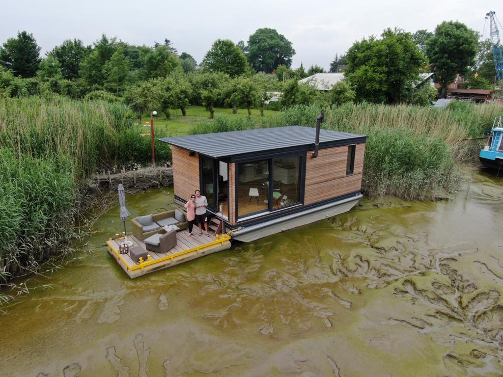 A houseboat is positioned on a tidal section of the river, surrounded by lush greenery and reeds. The modern design features large windows, offering views of the natural landscape. Outdoor seating is arranged on a floating terrace, enhancing the connection to the waterway.