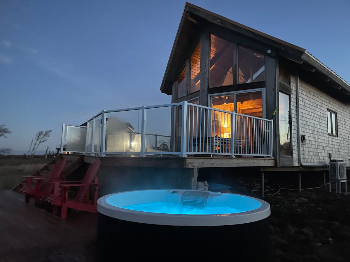 A hot tub with blue water is positioned in front of a modern cottage featuring floor-to-ceiling windows. The building is elevated and surrounded by a wooden deck, complemented by red outdoor chairs, offering a welcoming space for relaxation.