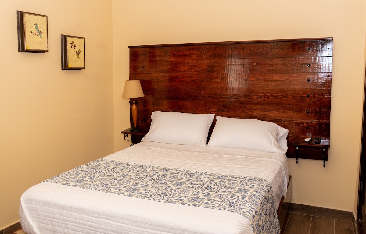 A cozy sleeping area features a natural wooden headboard with built-in shelves. Soft white bedding is complemented by a patterned blue throw. A bedside lamp softly illuminates the space, while framed artwork adds a decorative touch to the walls.