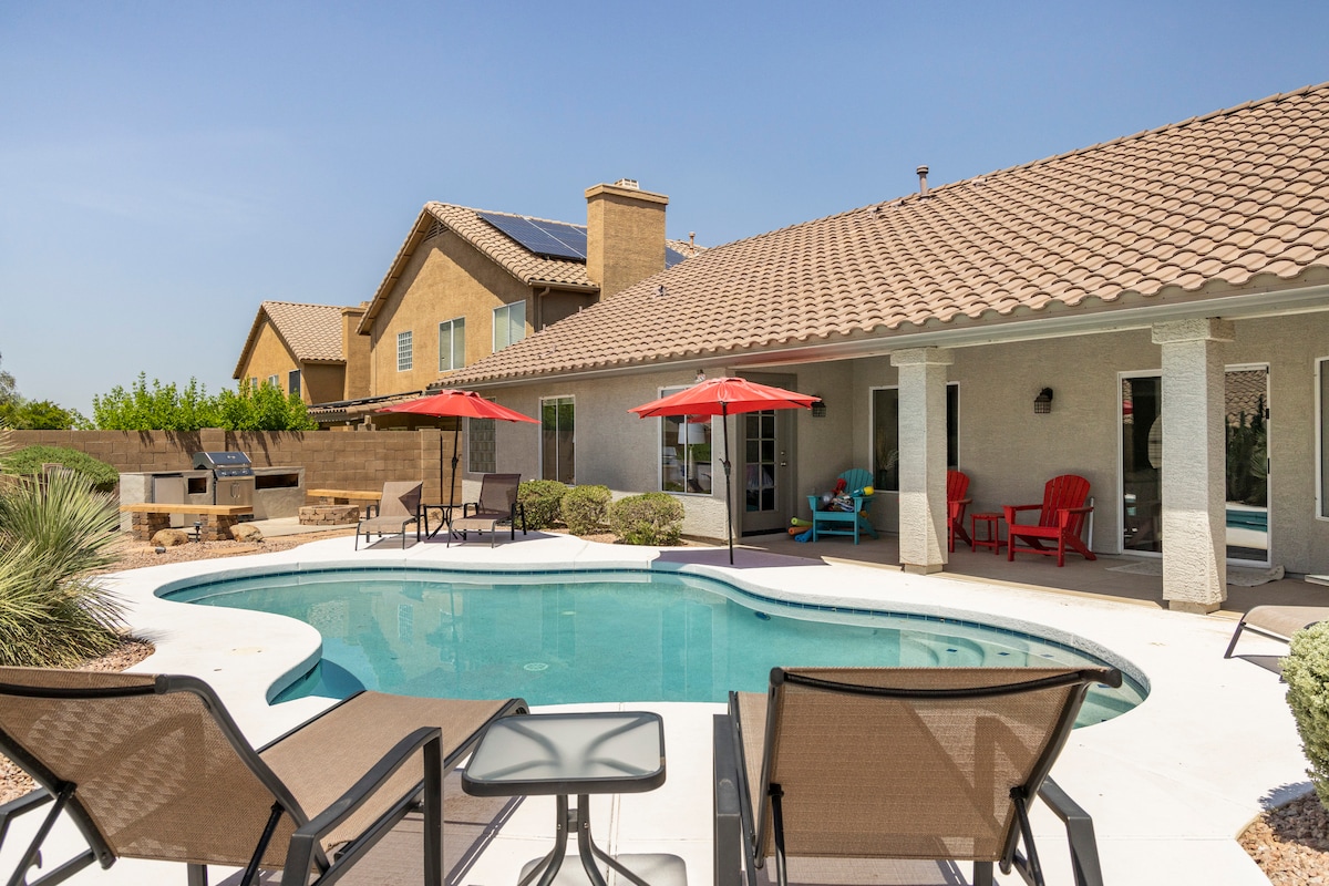A heated pool surrounded by lounge chairs and a small table invites relaxation. Colorful umbrellas provide shade, while a grill stands nearby. The home features a spacious covered patio, enhancing outdoor living with comfortable seating options and desert landscaping in the background.