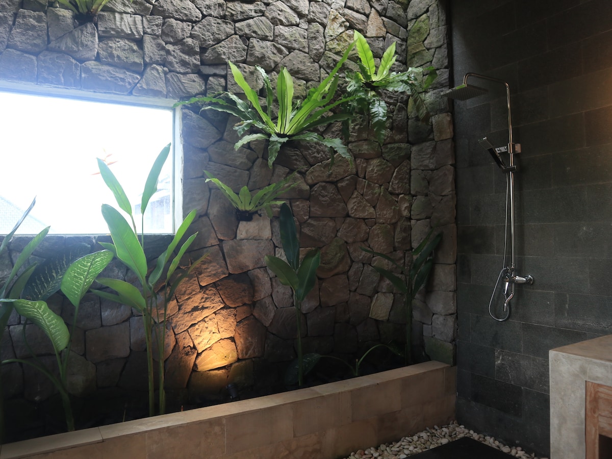 A semi-open bathroom features natural stone walls with vibrant green plants strategically placed for a calming atmosphere. A rainfall shower is mounted on one wall, while natural light filters in through a large window, highlighting the earthy tones and textures of the space.