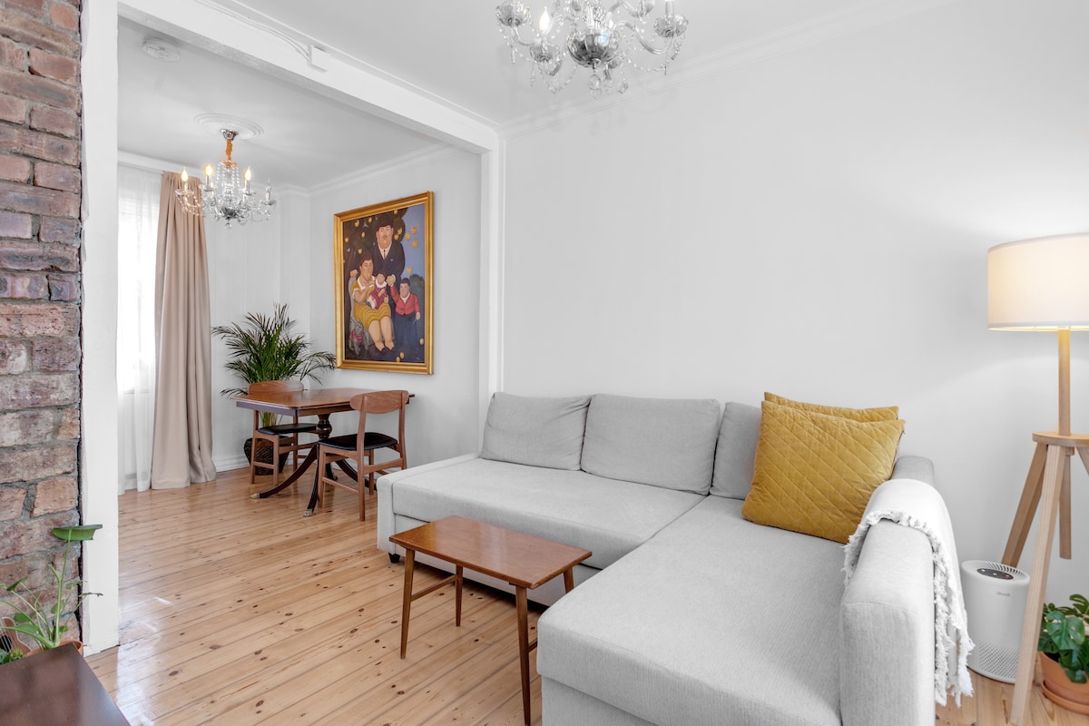 The living room features a modern gray sleeper sofa adorned with a yellow throw pillow. A pair of wooden tables are placed nearby, accompanied by a dining table set for two against a backdrop of bright walls and wood flooring. A colorful painting adds a vibrant focal point.