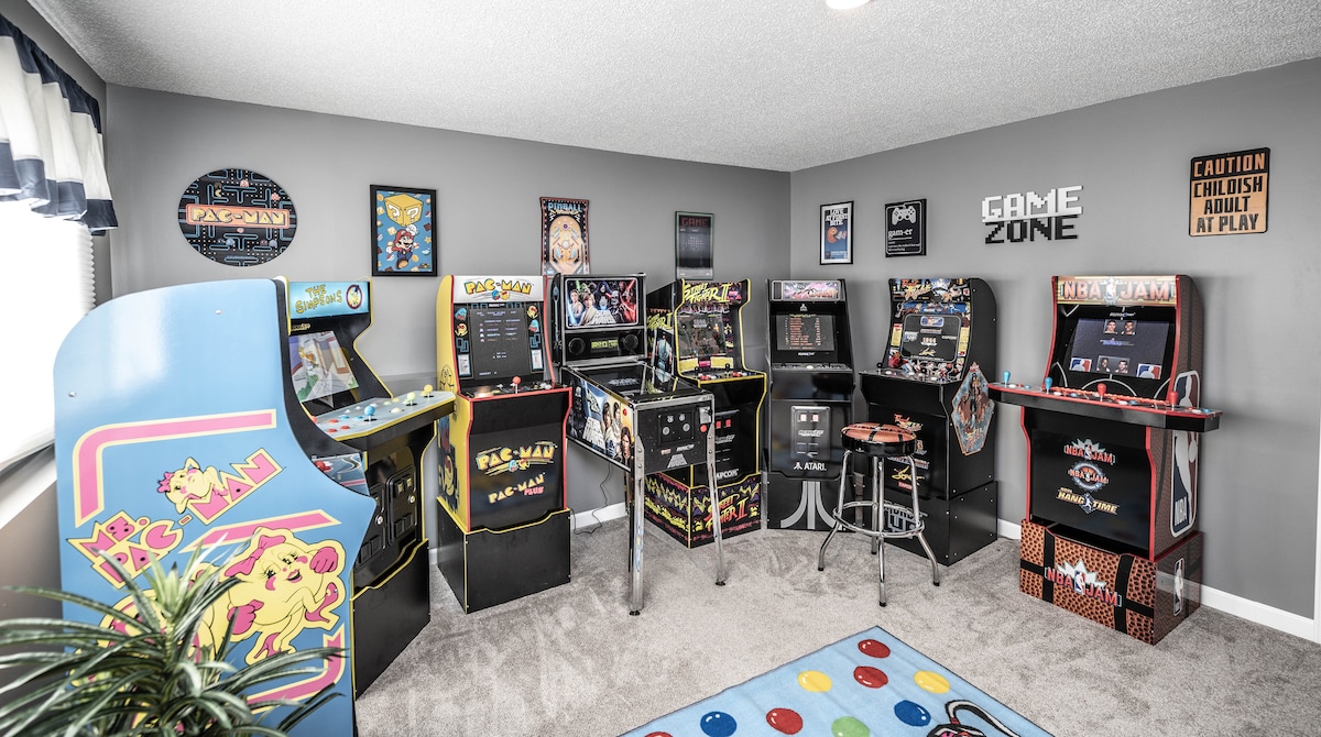 A vibrant arcade room is filled with classic game machines, including titles like Pac-Man and Ms. Pac-Man. The walls are adorned with game-themed decorations. A cozy rug with colorful dots lies on the carpeted floor, enhancing the playful ambiance of the space.
