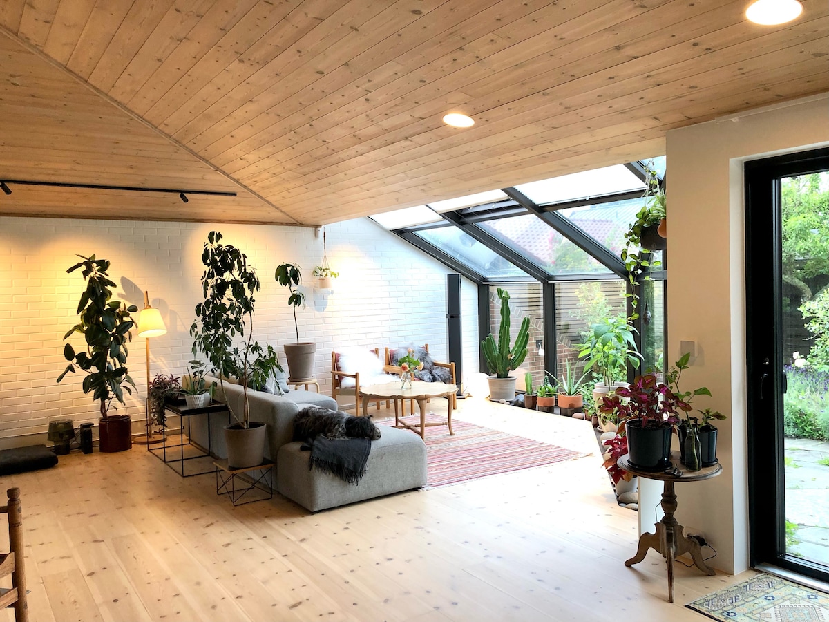 A bright and airy living space features a vaulted wooden ceiling and large glass walls, inviting natural light. Potted plants add greenery throughout the area. A cozy seating arrangement is highlighted by a wooden coffee table and decorative rugs, enhancing the connection to the outdoor garden.