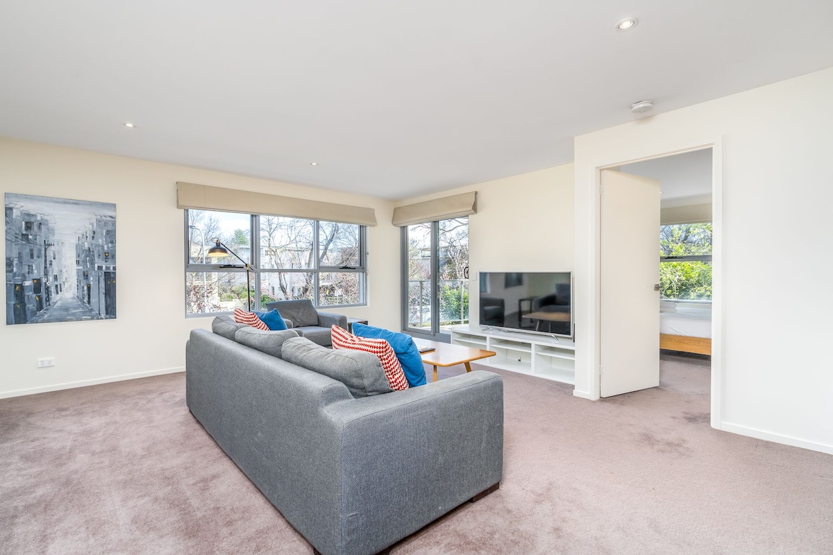The living room features a comfortable grey sofa with colorful cushions positioned near large windows that allow natural light to fill the space. A television is placed on a media unit opposite the seating area, with a visible doorway leading to another room.
