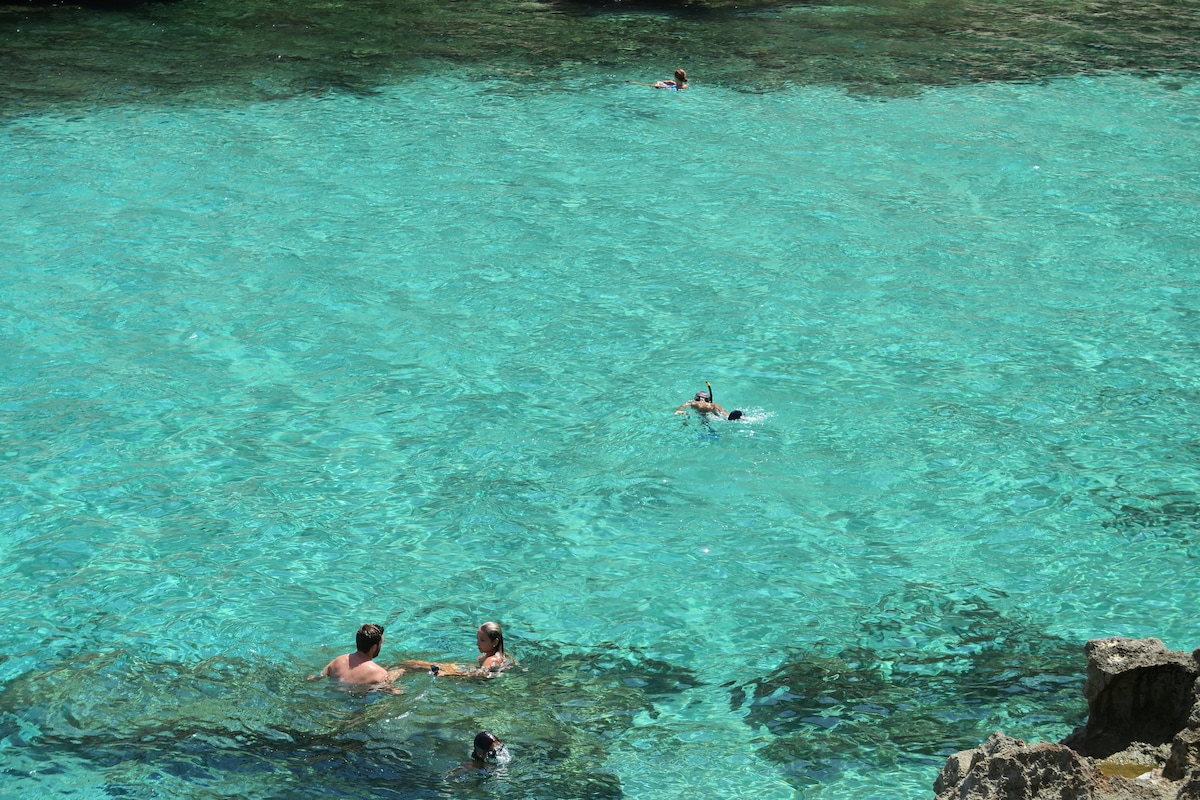 Clear turquoise waters are visible, with several individuals swimming and enjoying the refreshing environment. Rocky formations are scattered throughout the shallow areas, providing a natural landscape. The calm scene captures a pleasant day at the beach, ideal for relaxation and water activities.