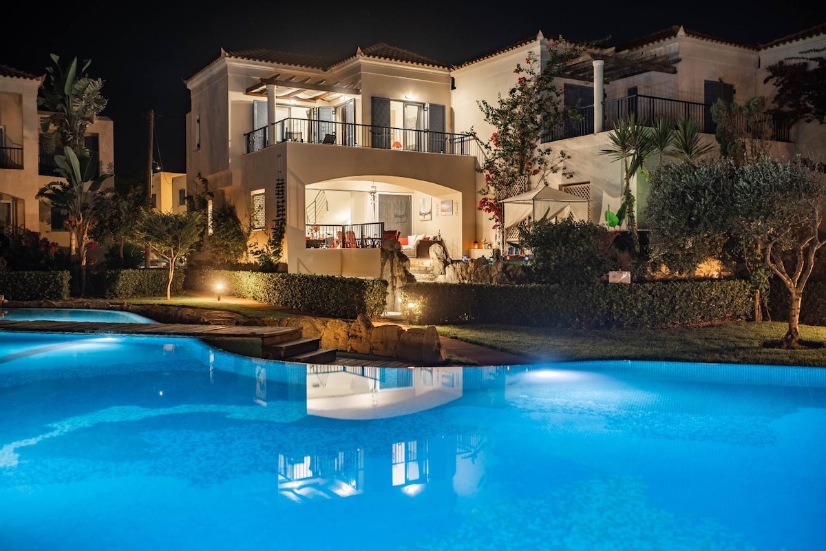 The exterior of Villa Rodo is illuminated at night, showcasing vibrant landscaping and a communal pool reflecting the glowing lights of the villa. Outdoor seating areas are visible on the porch, providing a welcoming environment for relaxation.