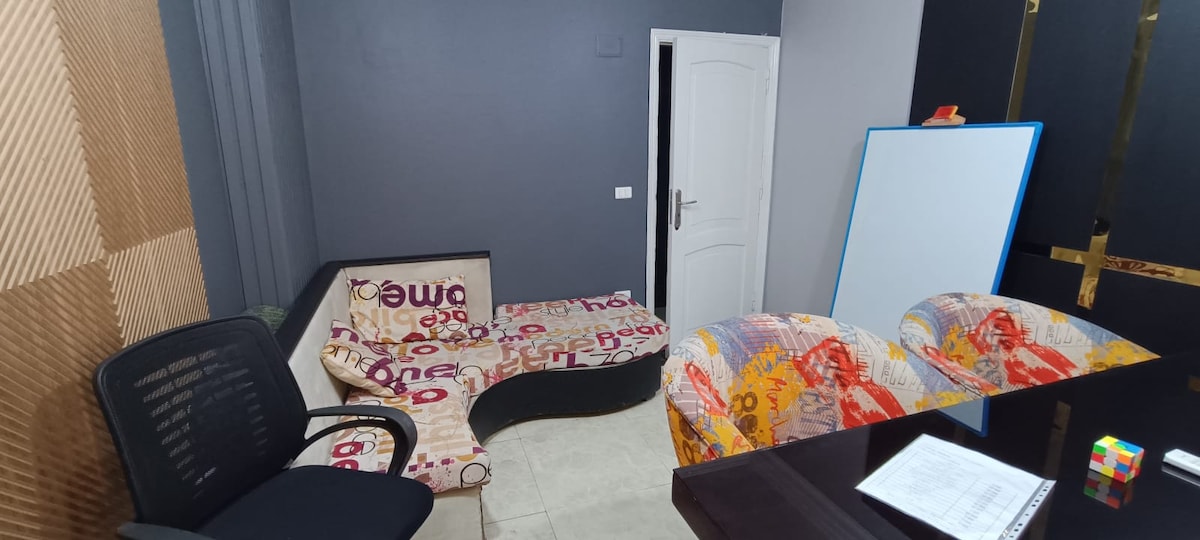 A cozy living space features a patterned seating area with a curved sofa and two chairs. A dark wooden table is positioned nearby, accompanied by a sheet of paper and a Rubik's cube. The walls are painted in soft gray tones, enhancing the room's minimalist aesthetic.