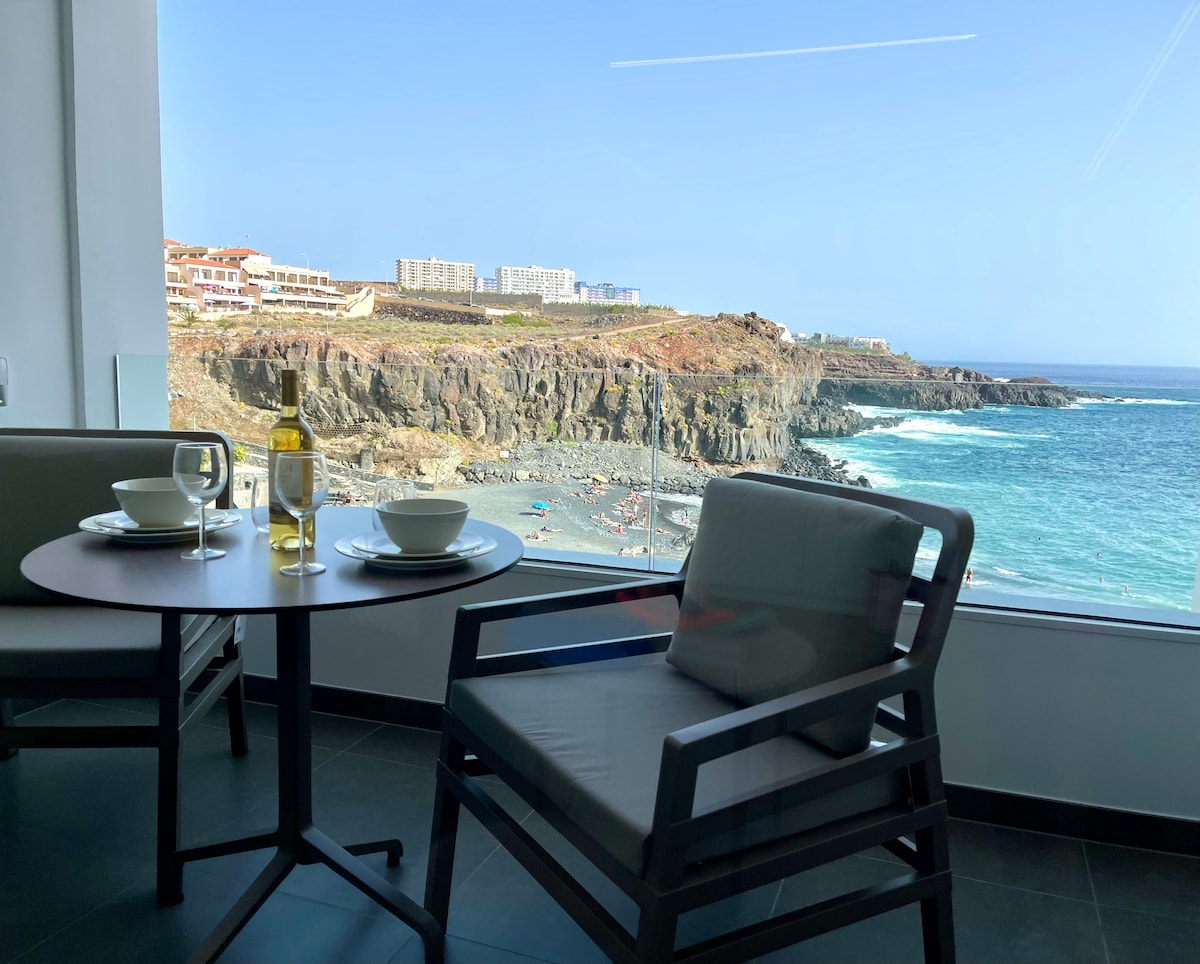 A dining area is positioned by a large window, revealing a scenic view of the rocky coastline and ocean. A round table is set with two plates, glasses, and a bottle of wine. The calm blue waters and sandy beach can be seen in the distance.