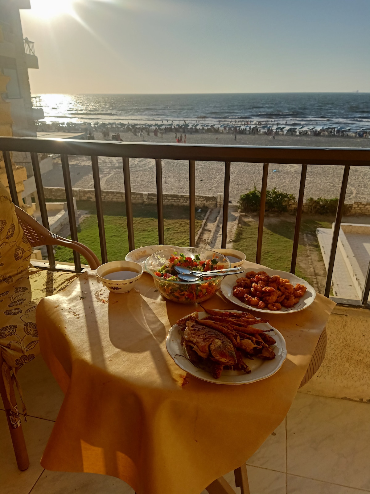 A balcony table is set with a variety of colorful dishes, including a salad, two plates of food, and drinks. In the background, the sunlit beach stretches along the horizon, while gentle waves meet the shore.