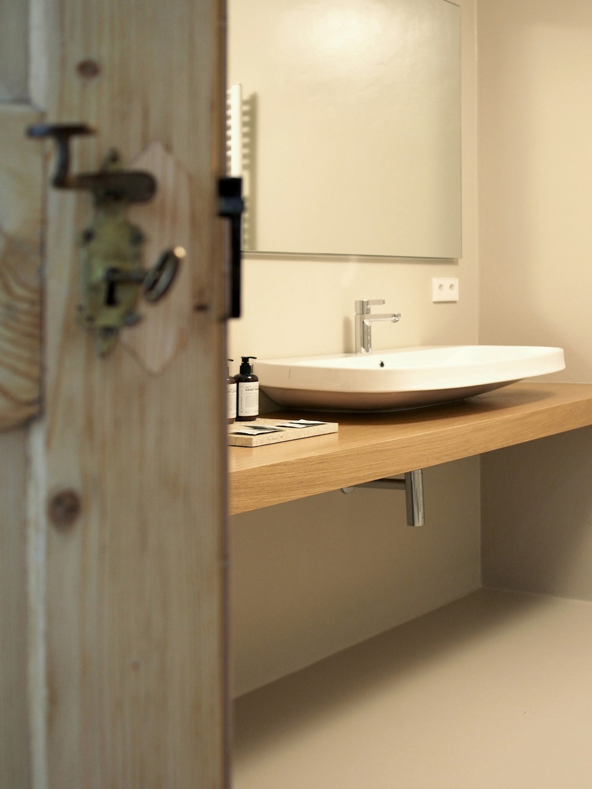 A modern bathroom setup is depicted, featuring a sleek wall-mounted sink on a wooden countertop. A large mirror reflects the minimalistic design. Toiletries are neatly arranged nearby. An intricate door handle adds a touch of elegance to the space, enhancing the overall tranquil feel.