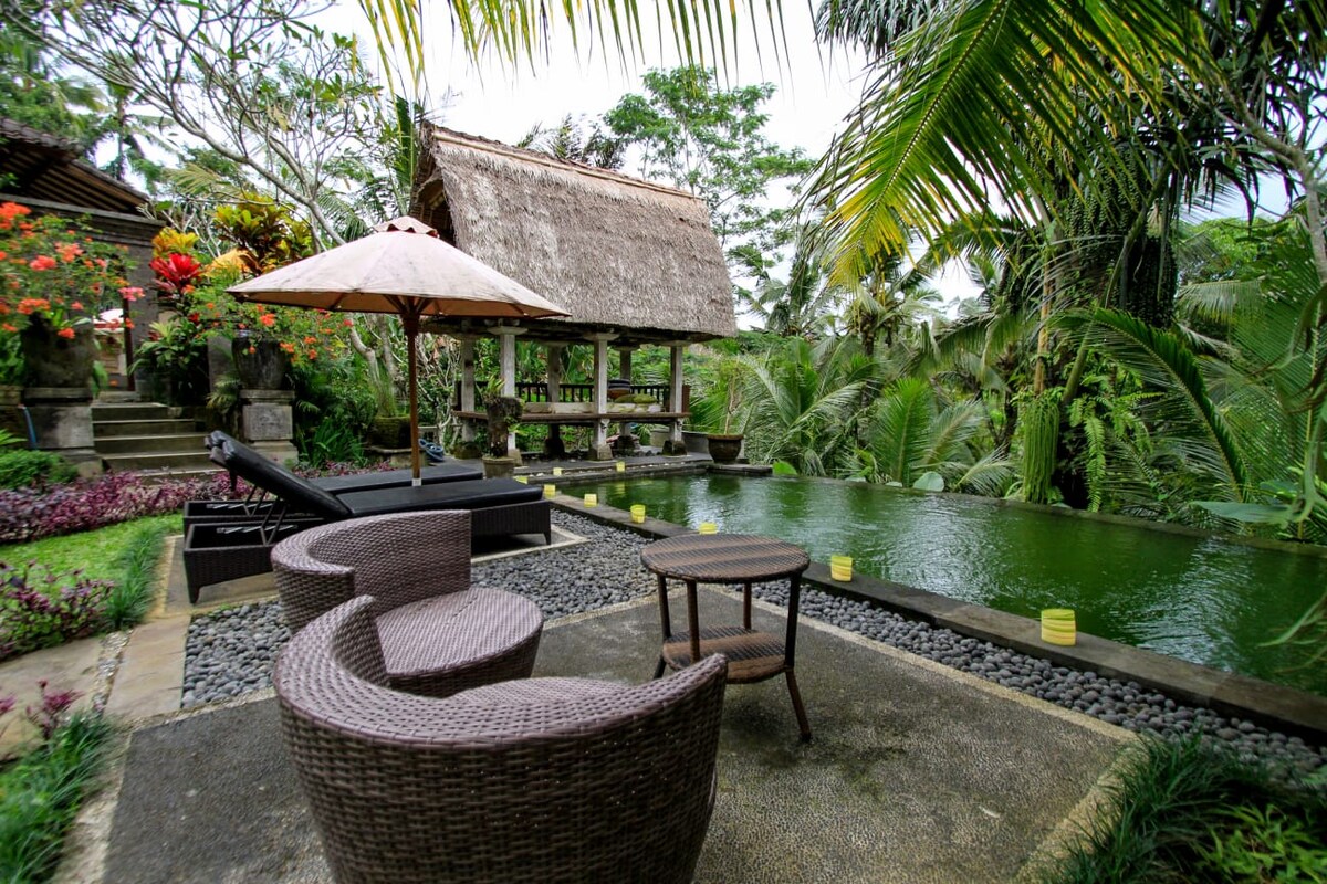 A serene outdoor lounging area features woven chairs and a round table, positioned next to a private swimming pool. Lush greenery surrounds the space, complemented by vibrant flowers and a thatched-roof pavilion in the background. Sun loungers are also present for relaxation.