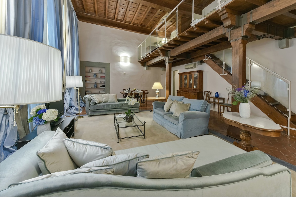 A spacious living area is highlighted by high wooden beams and large windows draped in soft blue curtains. Three comfortable sofas are arranged around a glass-topped coffee table. A dining table with chairs is visible in the background, along with stairs leading to an upper level.