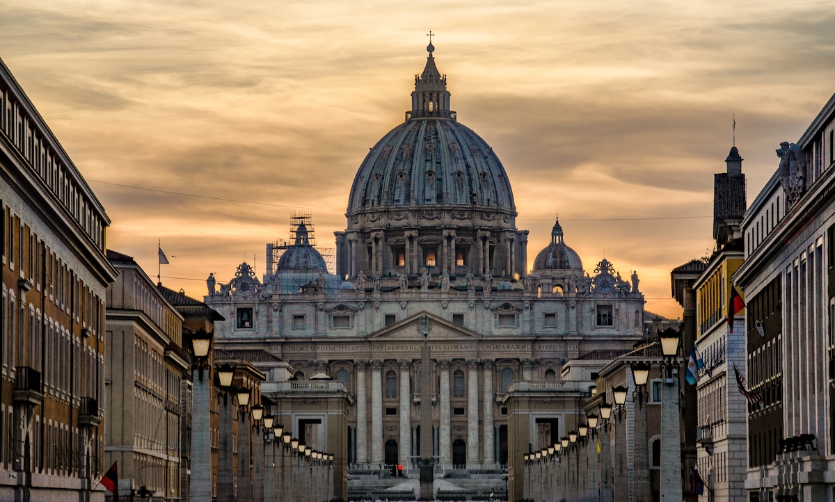 The majestic dome of St. Peter's Basilica is framed by elegant architecture on either side. The scene is set at dusk, with warm hues painting the sky, enhancing the grandeur of this iconic landmark.