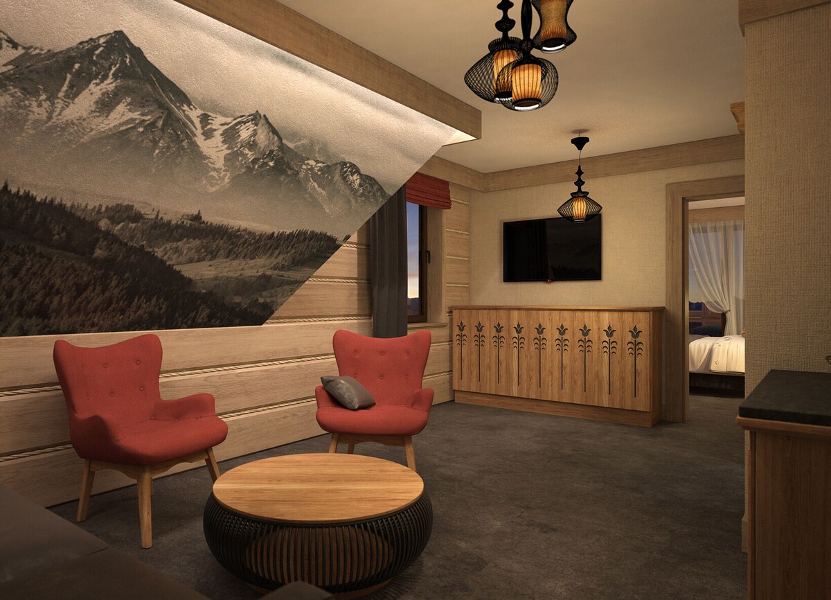 A spacious living area presents two red armchairs accompanied by a round wooden coffee table. A mountain mural adorns the wall, while stylish pendant lights hang from the ceiling. A flat-screen TV is mounted on the opposite wall, enhancing the modern alpine aesthetic.