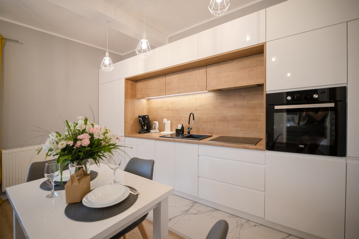 A modern kitchen area features white cabinetry with wooden accents and black appliances. A dining table with grey chairs is set for a meal, accompanied by fresh flowers. Soft lighting is provided by pendant fixtures overhead, enhancing the contemporary design.