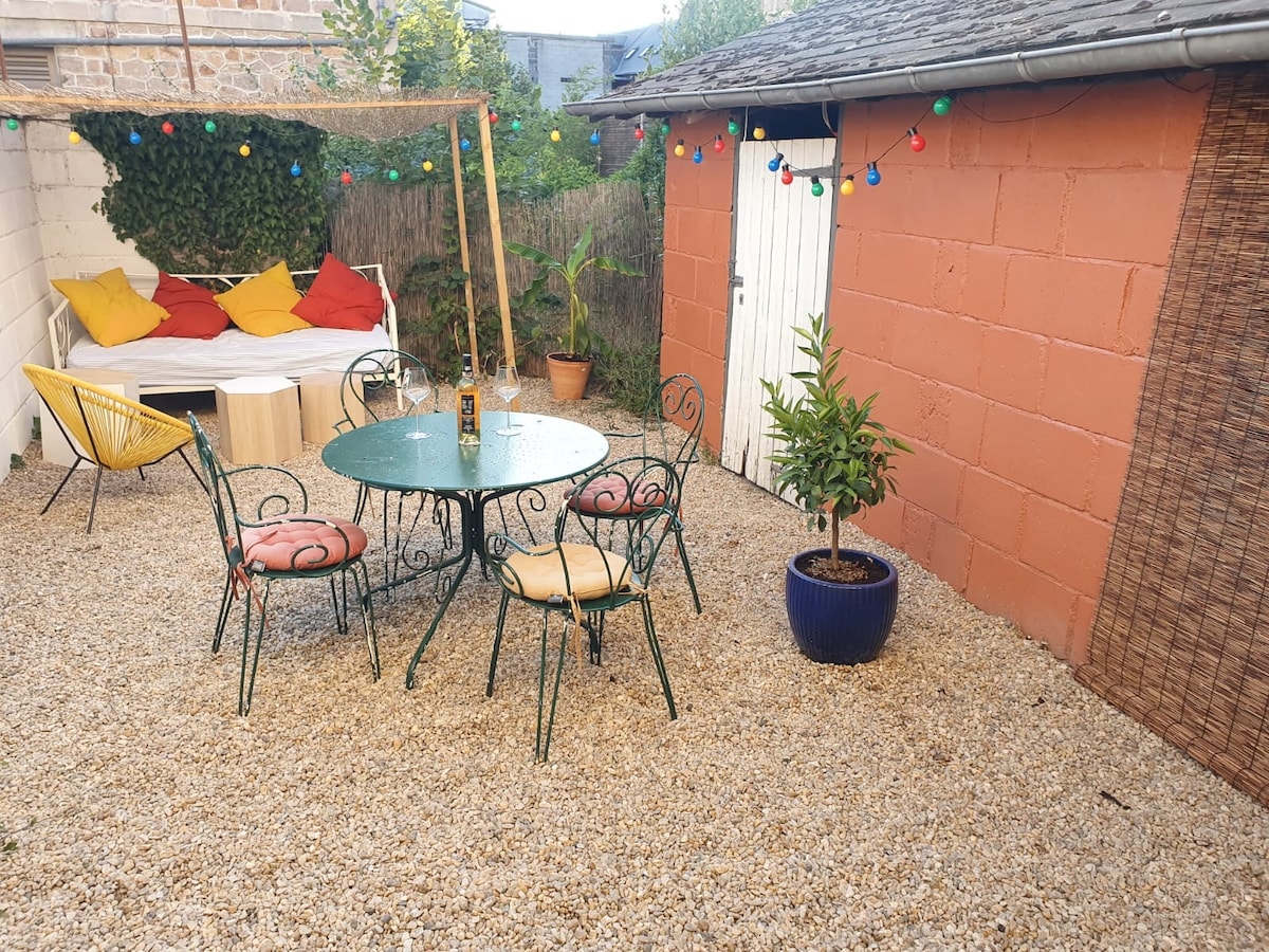 An outdoor space features a gravel flooring with a round metal dining table surrounded by six chairs. A lounge area with colorful cushions is visible, along with a potted plant and a small storage shed. String lights hang above, creating a cozy atmosphere.