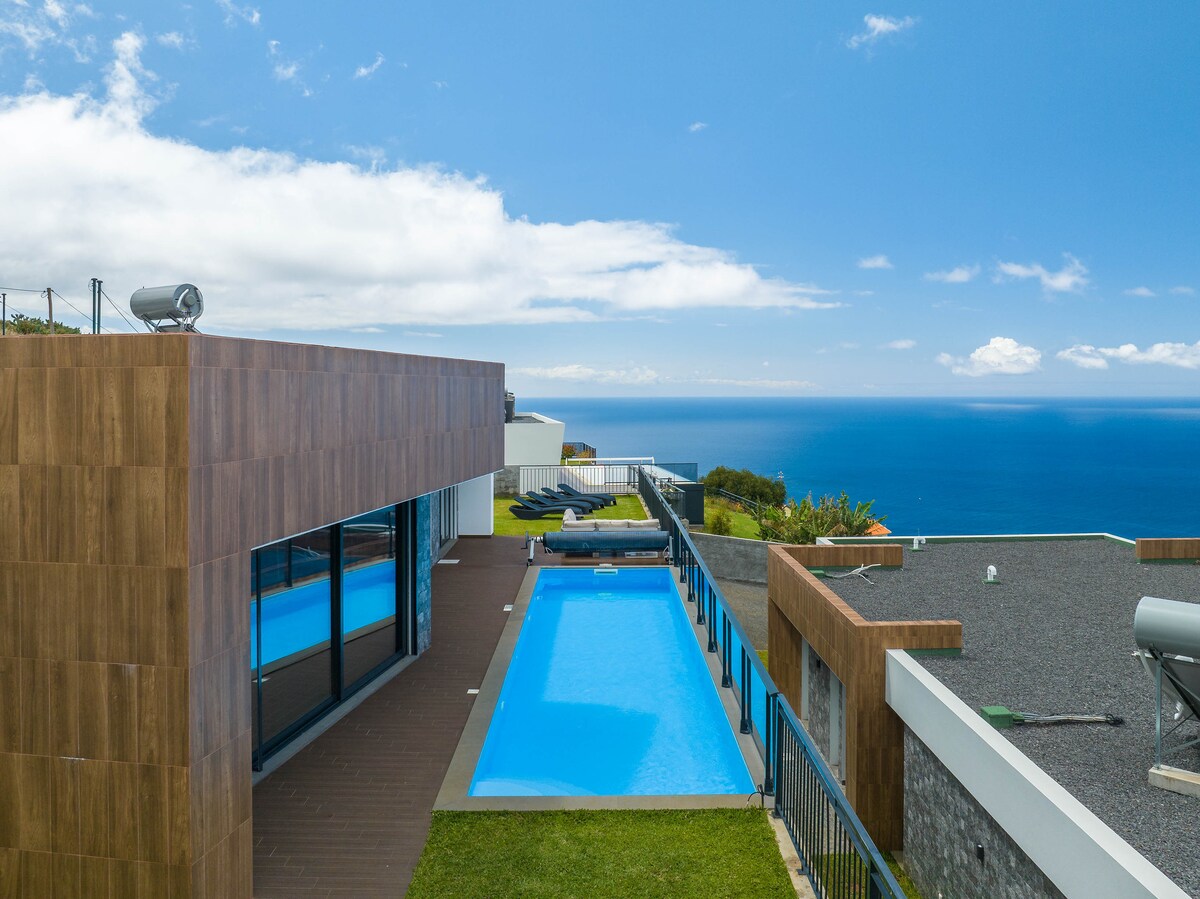 A private swimming pool is situated adjacent to the modern house, with expansive glass windows offering views of the ocean. The area is surrounded by well-maintained grass and features lounge chairs positioned for relaxation, all under a clear blue sky.