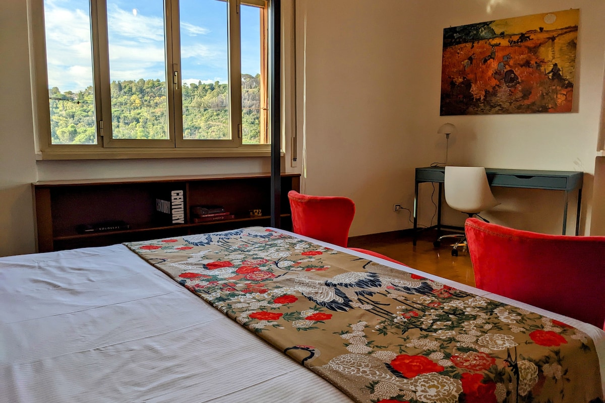 A bright bedroom is featured with large windows offering a scenic view of greenery. A spacious bed is adorned with a decorative coverlet, while a desk and chair set is positioned nearby. A colorful artwork hangs on the wall, adding to the room's character.